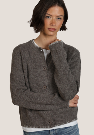 A woman wearing a dark brown heathered knit cardigan sweater with button-front closure and ribbed trim, paired with a white top and blue jeans, against a plain light beige studio background.