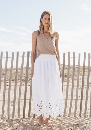 Dylans Clothing. Woman in beige top and white skirt standing on a sandy beach with a wooden fence in the background.