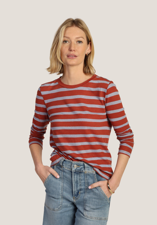 A woman modeling a long-sleeve crewneck top with horizontal rust red and light gray stripes, paired with light-wash blue jeans, against a solid light gray background.