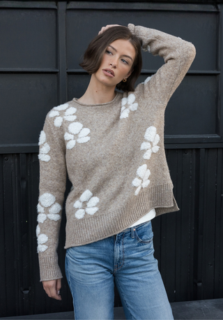 A woman wearing a beige knit sweater with large white floral motifs and blue jeans, posing against a dark paneled wall.
