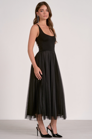 A woman modeling a sleeveless black midi dress with a fitted bodice and a flowing tulle skirt, paired with black high heels, against a plain light gray studio background.