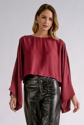 A woman modeling a cropped, long-sleeved blouse in deep burgundy satin with wide draped sleeves, paired with black faux-leather pants, against a light gray studio background.