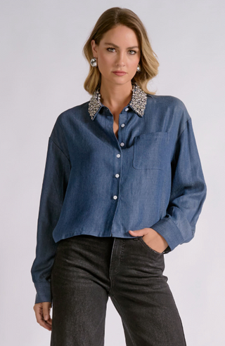 A woman modeling a blue long-sleeve button-up shirt with a decorative silver and black patterned collar, paired with dark gray jeans, against a light gray studio background.