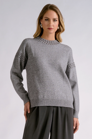 A woman modeling a heather gray knit sweater with decorative silver-toned embroidery around the crew neck and on the upper sleeves, paired with dark gray wide-leg pants, against a light gray studio background.