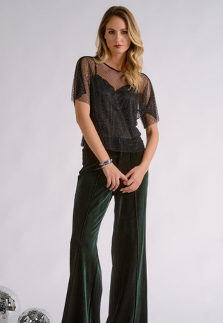 A woman modeling a black sheer mesh short-sleeve top with a black lace camisole underneath, paired with dark green wide-leg trousers, against a light gray studio background.