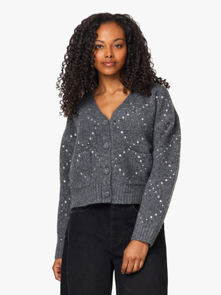 A woman with curly dark hair wearing a dark gray knit cardigan with white embroidered star patterns and black pants, photographed against a solid light gray background.