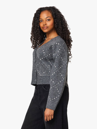A woman with long curly black hair is shown from the side, wearing a gray knit cardigan with white sequins and black pants against a plain white studio background.