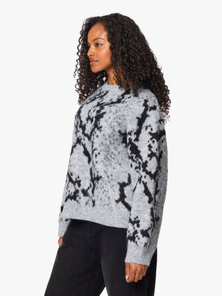 A woman with curly dark hair wearing a gray and black snakeskin-patterned knit sweater and black pants, shown in a side-profile view against a plain light gray studio background.