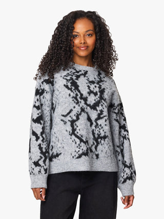 A woman with curly dark hair wearing a light gray knit sweater with a black abstract pattern, paired with black pants, against a plain light gray background.