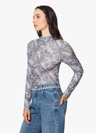 A woman modeling a long-sleeve mock neck top with a gray and white snakeskin pattern, paired with blue jeans, against a plain white background.