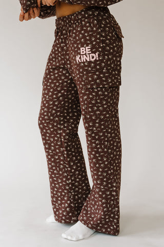 Brown flared pajama pants with a white floral pattern and the words 'BE KIND!' printed on the left thigh, shown on a model against a light gray background.