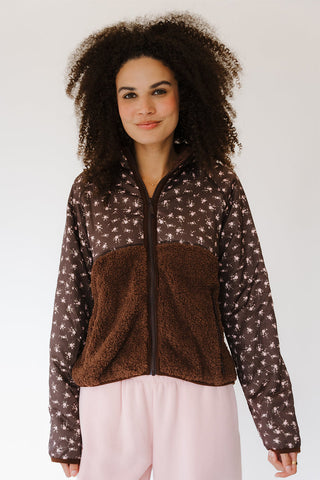 A woman with curly dark hair wearing a brown fleece zip-up jacket with a patterned upper section and light pink pants, photographed against a plain light gray studio background.