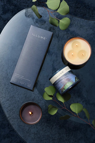 A set of Illume candles from the Hidden Lake Collection, displayed on a dark surface with green leaf accents. The candles are in various sizes, with one lit to showcase the flame.