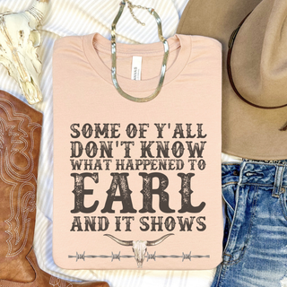 A folded light peach t-shirt with a western-themed graphic print reading 'SOME OF 'Y'ALL DON'T KNOW WHAT HAPPENED TO EARL AND IT SHOWS' with a skull and barbed wire design. The shirt is styled in a flat lay with a silver necklace, a cowboy boot, a hat, and blue jeans.
