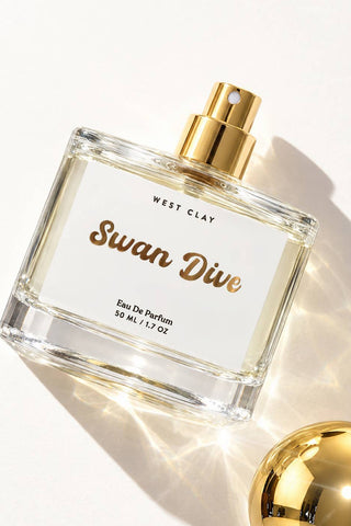 West Clay Swan Dive Eau De Parfum 95ml glass bottle with a gold spray cap and a white label, styled on a white background with a gold reflective sphere prop.