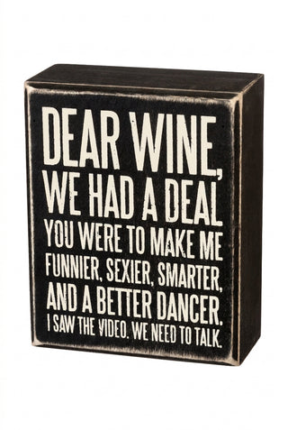 A rectangular black decorative block sign with distressed white text that reads, 'DEAR WINE, WE HAD A DEAL YOU WERE TO MAKE ME FUNNIER, SEXIER, SMARTER, AND A BETTER DANCER. I SAW THE VIDEO, WE NEED TO TALK.