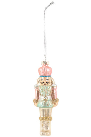 A glass nutcracker Christmas ornament with a pastel pink crown, mint green dress, and gold glitter accents, hanging from a silver string on a white background.