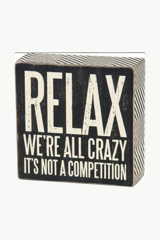 Black wooden block with white text 'RELAX WE'RE ALL CRAZY IT'S NOT A COMPETITION' on a white background