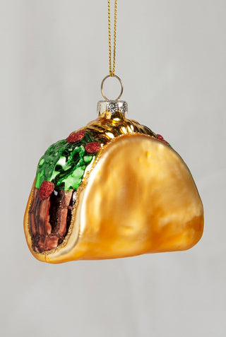 A novelty Christmas ornament shaped like a taco, with a glossy gold shell, green lettuce, red tomatoes, and brown meat filling, hanging by a gold string against a light gray background.